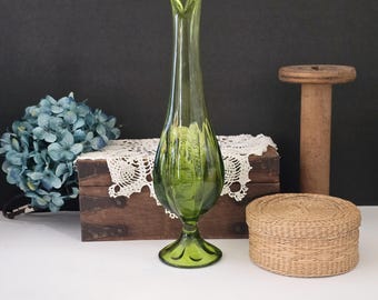 May include: A tall, green glass vase with a flared opening and a fluted design. The vase has a round base and is sitting on a wooden surface.