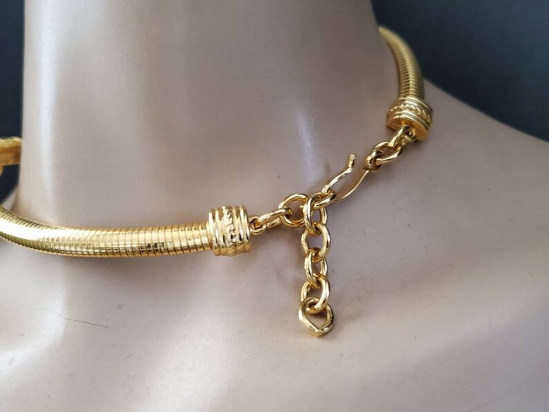 May include: A gold-toned necklace with a textured, ribbed design. The necklace is displayed on a mannequin and features a chain and clasp closure. The jewelry has a bright, shiny finish.