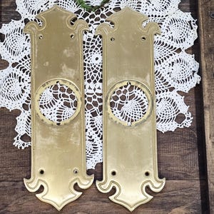 May include: Two antique brass door plates with ornate detailing. Each plate has a rectangular shape with a decorative top and bottom, and a circular cutout in the center. The plates are set against a white lace doily on a wooden surface.