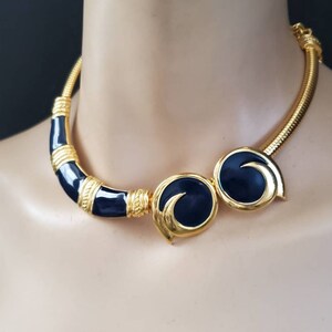 May include: Gold-tone necklace with navy blue enamel accents. The necklace features a curved bar and two circular elements with a swirl design. The necklace has a textured, snake-like chain.