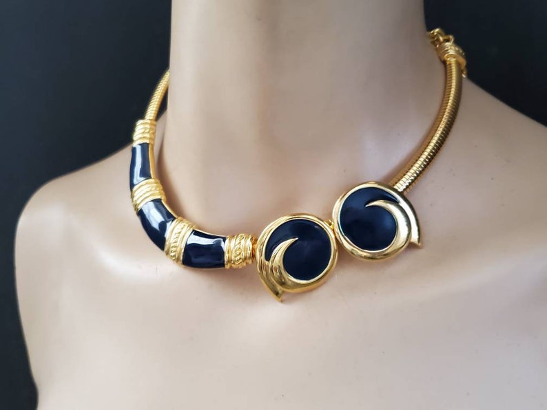 May include: Gold-tone necklace with navy blue enamel accents. The necklace features a curved bar and two circular elements with a swirl design. The necklace has a textured, snake-like chain.