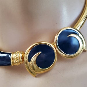 May include: A gold-toned necklace with navy blue enamel accents. The necklace features a coiled chain and circular, stylized elements. The design includes a combination of smooth and textured surfaces, creating a striking contrast.