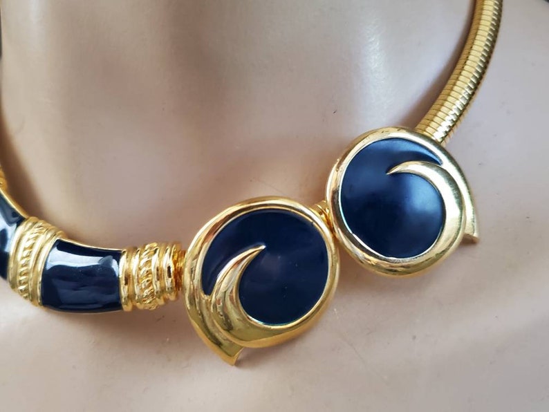 May include: A gold-toned necklace with navy blue enamel accents. The necklace features a coiled chain and circular, stylized elements. The design includes a combination of smooth and textured surfaces, creating a striking contrast.