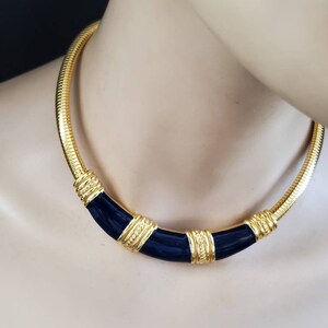 May include: A gold-toned necklace with a snake chain design. The necklace features a curved, navy blue enamel center section with gold accents. The necklace is displayed on a mannequin.