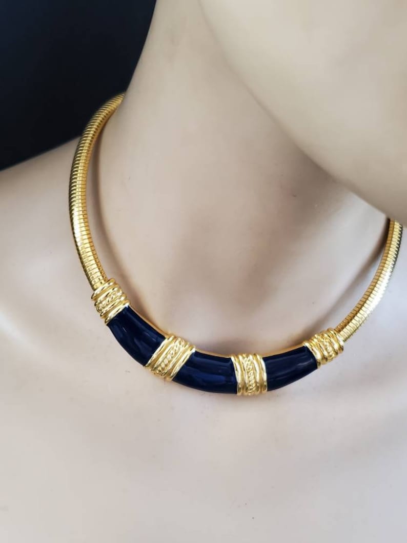 May include: A gold-toned necklace with a snake chain design. The necklace features a curved, navy blue enamel center section with gold accents. The necklace is displayed on a mannequin.