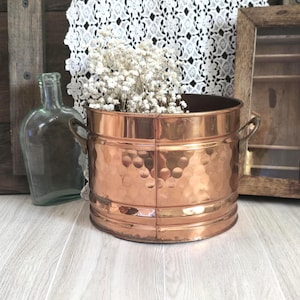 May include: A copper-colored metal bucket with a hammered texture, holding white flowers. The bucket has two small handles and is set against a backdrop of a lace doily, a glass bottle, and a wooden frame.