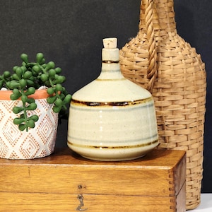 May include: A ceramic bottle with a cork stopper, featuring a beige body with brown stripes. A woven bottle and a small potted succulent sit behind the bottle. The items are on a wooden box.