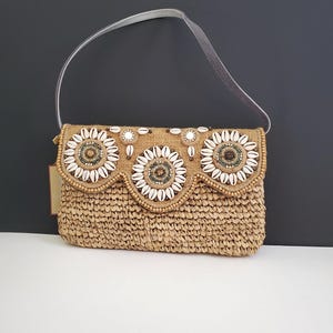 May include: A tan straw clutch bag with a scalloped edge and three circular embellishments made of white seashells, brown beads, and gold-toned accents. The bag has a light gray leather strap.