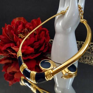 May include: A gold-toned necklace and matching earrings set with navy blue enamel accents. The necklace has a textured, snake-like chain and a curved design. The earrings are round with a navy blue center. The jewelry is displayed on a white hand-shaped stand.