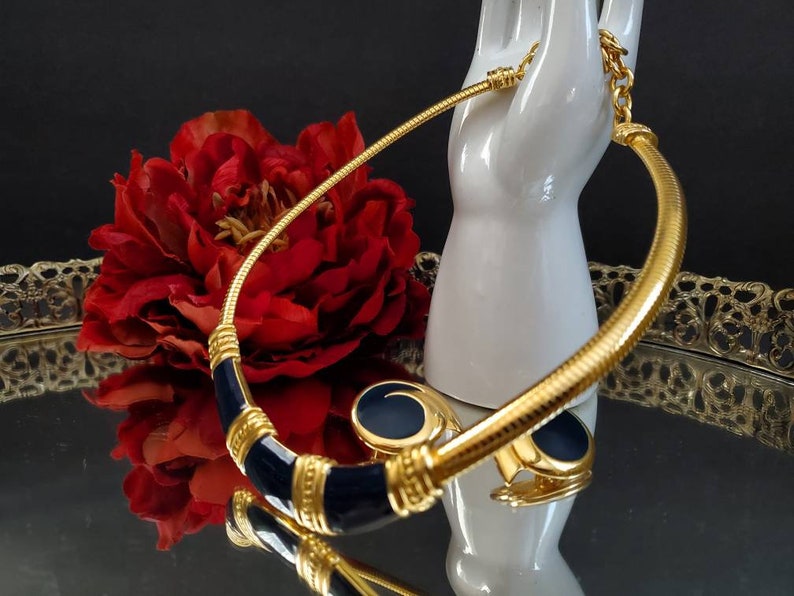 May include: A gold-toned necklace and matching earrings set with navy blue enamel accents. The necklace has a textured, snake-like chain and a curved design. The earrings are round with a navy blue center. The jewelry is displayed on a white hand-shaped stand.