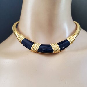 May include: A gold-tone choker necklace with a navy blue enamel center and gold accents. The necklace has a ribbed design and is displayed on a mannequin. The necklace is a statement piece.