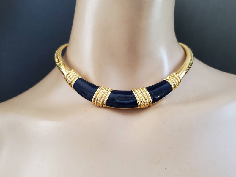 May include: A gold-tone choker necklace with a navy blue enamel center and gold accents. The necklace has a ribbed design and is displayed on a mannequin. The necklace is a statement piece.