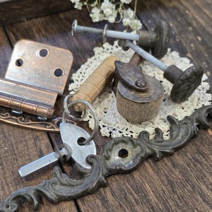 May include: An assortment of vintage hardware components, including metal drawer pulls, hinges, and keys, arranged on a wooden surface. The hardware pieces are in various shades of bronze, copper, and silver, with a few wooden elements.