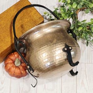 May include: A hammered brass cauldron with a wide rim and a rounded body. It features a black metal swing handle attached to the sides and three black metal feet at the base. The surface of the brass has a textured, dimpled finish throughout.