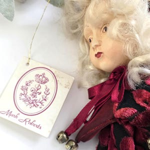May include: A decorative doll with a porcelain face and blonde curly hair. The doll wears a burgundy and black patterned outfit with small silver bells. A tag with the words "Mark Roberts" is attached.