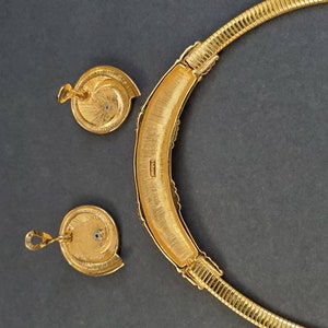 May include: A gold-toned necklace and matching earrings set. The necklace is a rigid collar with a textured, ribbed design. The earrings are round with a spiral design. The set is displayed on a dark gray background.