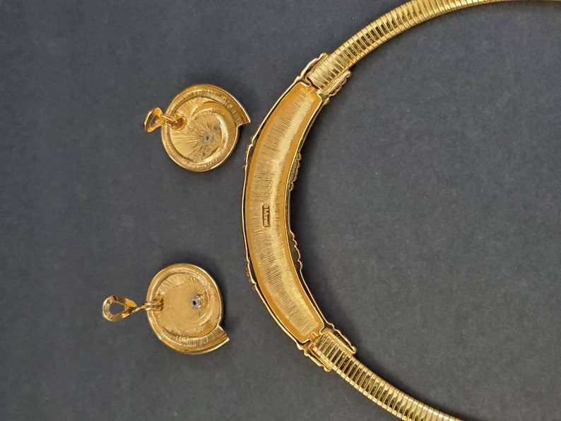 May include: A gold-toned necklace and matching earrings set. The necklace is a rigid collar with a textured, ribbed design. The earrings are round with a spiral design. The set is displayed on a dark gray background.
