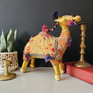 May include: A colorful, handmade camel figurine with gold trim, purple, pink, and green accents. The camel is standing on a white surface with a brass candlestick and a book.