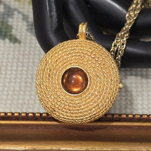May include: A gold-toned pendant necklace with a circular, woven design. The pendant features a central, amber-colored stone. The necklace chain is also gold-toned. The jewelry is displayed against a black background.