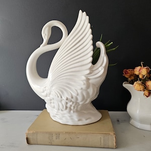 May include: White ceramic swan vase with a detailed design. The swan's wings are spread out and the neck is curved into a circle. The vase is sitting on a vintage book.