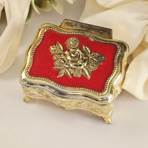 May include: A small, ornate jewelry box with a red textured top and gold-colored trim. The top features a gold floral design. The box has small feet and a hinged lid. The overall design is vintage and decorative.