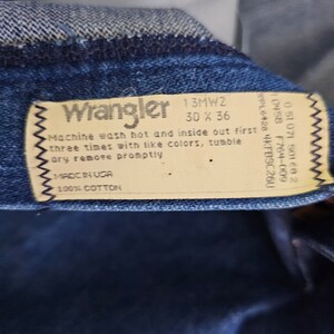 May include: A white care label with black text for Wrangler jeans. The label says "Machine wash hot and inside out first three times with like colors, tumble dry remove promptly. Made in USA. 100% Cotton."