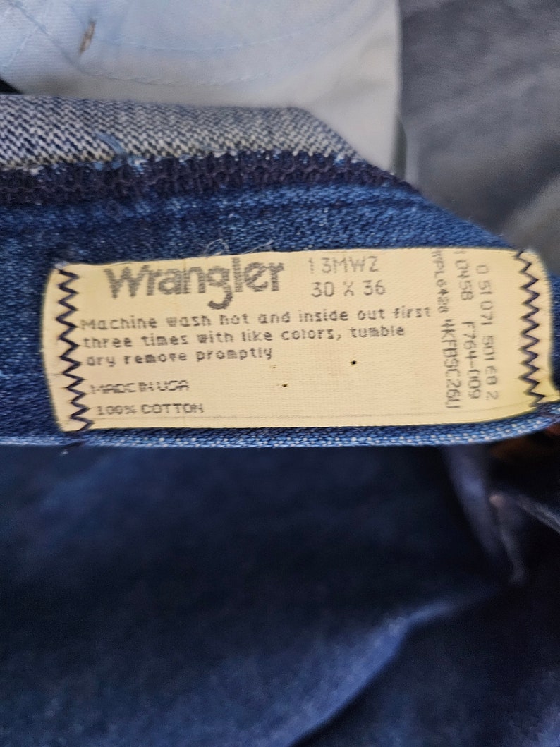 May include: A white care label with black text for Wrangler jeans. The label says "Machine wash hot and inside out first three times with like colors, tumble dry remove promptly. Made in USA. 100% Cotton."