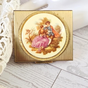 May include: A vintage gold-toned compact with a circular, cream-colored lid featuring a romantic scene of two figures in period clothing. The lid is framed with a gold rope-like border. The compact has a rectangular shape.