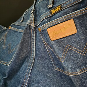 May include: A pair of blue denim jeans with a brown leather patch that says "Wrangler" on the back pocket. The jeans have a classic five-pocket style and a straight leg.