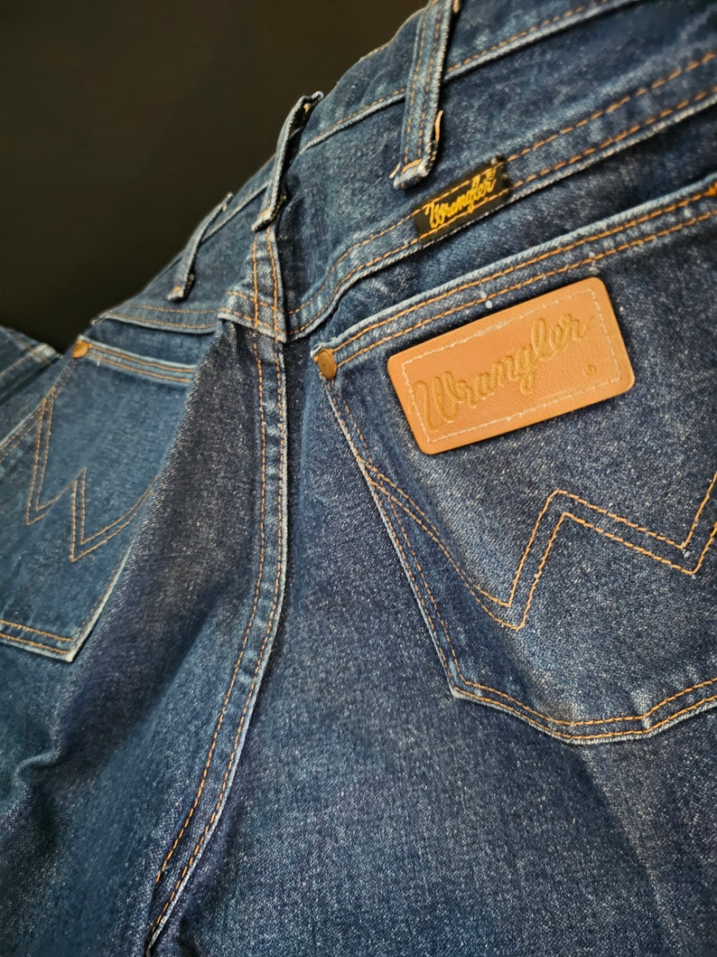 May include: A pair of blue denim jeans with a brown leather patch that says "Wrangler" on the back pocket. The jeans have a classic five-pocket style and a straight leg.