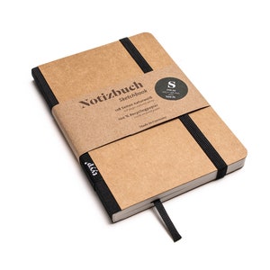 Op de afbeelding: Een bruin notitieboek met een zwart elastiek en een zwart label met de tekst "Notizbuch Sketchbook 128 Seiten naturweiß 100 % Recyclingpapier Made in Germany". Het label heeft ook een kleine zwarte cirkel met de letter "S" erin.