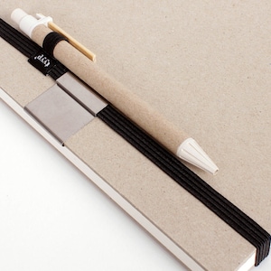 Puede incluir: Un cuaderno marrón con una banda elástica negra y un bolígrafo beige con una punta blanca. El bolígrafo se mantiene en su lugar mediante un clip de metal plateado.