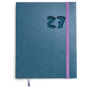 Könnte beinhalten: Ein rechteckiges blaues Notizbuch mit texturiertem Einband und einer ausgestanzten Zahl 27 auf der oberen rechten Seite. Das Notizbuch wird durch ein vertikales violettes Gummiband verschlossen und hat ein passendes violettes Lesebändchen.