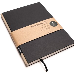Pode incluir: Um caderno de esboços preto com uma faixa de papel castanho à volta. A faixa tem o texto "Notizbuch Sketchbook 128 Seiten naturweiß 100% Recyclingpapier Made in Germany" impresso nela. O caderno de esboços tem um marcador de fita preto.