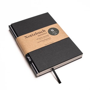 Op de afbeelding: Een zwart schetsboek met een kraftpapieren band met de tekst "Notizbuch Sketchbook". Het notitieboek heeft een zwart lint als bladwijzer en een gestreepte rand. De kaft is gemaakt van een textuur.