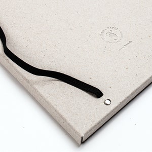 以下が含まれることがあります： 黒いゴムバンドで留める、薄いグレーのノート。表紙には「DESIGN & PAPER」の文字と小さなロゴがエンボス加工で施されています。ゴムバンドの穴は金属製のグロメットで補強されています。