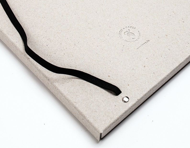 以下が含まれることがあります： 黒いゴムバンドで留める、薄いグレーのノート。表紙には「DESIGN & PAPER」の文字と小さなロゴがエンボス加工で施されています。ゴムバンドの穴は金属製のグロメットで補強されています。