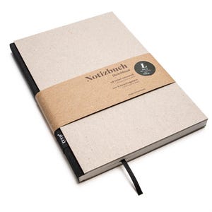 Puede incluir: Un cuaderno de bocetos rectangular con una cubierta de color beige claro y un lomo negro. La cubierta está envuelta con una banda de papel marrón que dice "Notizbuch Sketchbook". Una cinta negra como marcador de libro es visible. El cuaderno está etiquetado como "L".