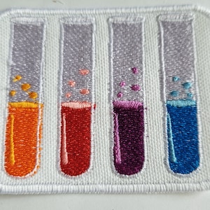 May include: Four glass test tubes with different colored liquids inside. The liquids are orange, red, purple, and blue. Each tube has small white dots inside.
