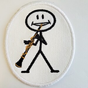 May include: White embroidered patch with a stick figure playing a clarinet. The stick figure has a smiling face and is holding the clarinet in its right hand.