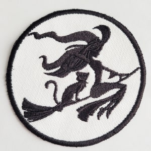 Könnte beinhalten: Ein weißer runder Patch mit schwarzer Stickerei einer Hexe, die auf einem Besen mit einer schwarzen Katze fliegt.