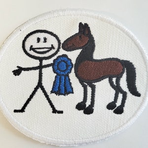 May include: Embroidered patch featuring a stick figure with a smiling face, holding a blue ribbon next to a brown and black horse. The patch is round with a white background and a stitched border, suitable for equestrian enthusiasts.