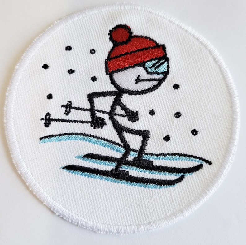 May include: Embroidered patch of a stick figure skier wearing a red hat and goggles, skiing down a snowy slope. The patch is circular and has a white background.