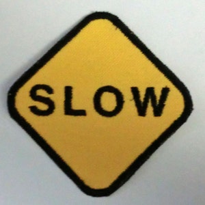 Könnte beinhalten: Ein gelbes, rautenförmiges Schild mit schwarzem Rand und dem Wort "SLOW" in schwarzen Buchstaben.