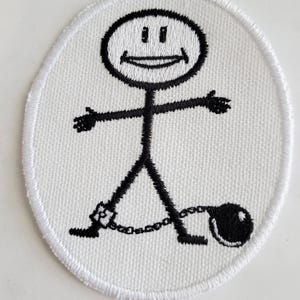 Puede incluir: Parche blanco bordado con una figura de palo que lleva una cadena y una bola y cadena. La figura de palo tiene una cara sonriente.