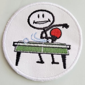 Könnte beinhalten: Ein weißer gestickter Aufnäher mit einer Strichmännchenfigur, die Tischtennis spielt. Die Strichmännchenfigur lächelt und hält einen roten Schläger. Der Tischtennistisch ist grün mit einem weißen Netz. Der Aufnäher ist rund und hat einen weißen Rand.