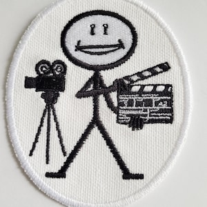 Könnte beinhalten: Weißer gestickter Aufnäher mit einer Strichmännchenfigur, die eine Filmklappe und eine Kamera auf einem Stativ hält. Die Strichmännchenfigur lächelt mit zwei Punkten für Augen.