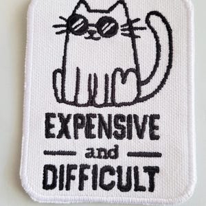 Op de afbeelding: Witte geborduurde patch met een zwarte lijntekening van een kat met een zonnebril. De tekst "EXPENSIVE and DIFFICULT" staat onder de kat.