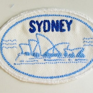 May include: A white oval patch with blue stitching around the edges. The patch features a blue embroidered image of the Sydney Opera House and the word "SYDNEY" in blue embroidery.