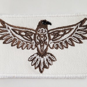 Könnte beinhalten: Ein gestickter Aufnäher mit einem stilisierten Adler-Design in Braun auf weißem Hintergrund.